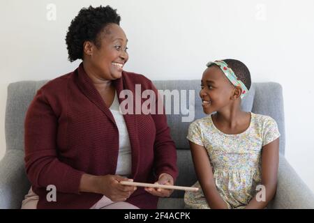Glückliche ältere afroamerikanische Großmutter, die neben lächelnd auf der Couch sitzt Enkelin mit Tablet Stockfoto