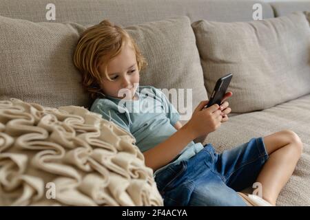 Kaukasischer Junge mit langen blonden Haaren auf Couch sitzend Smartphone Stockfoto