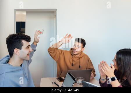 Die drei Freunde arbeiten zu Hause zusammen. Glückliche Männer geben einen High-Five, während das Weibchen sie ansieht Stockfoto