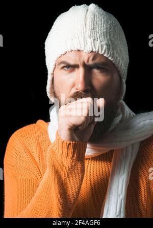 Mann hustet. Kranker Mann hustet in seine Faust. Bärtig schöner Kerl in Pullover, Hut und Schal. Stockfoto