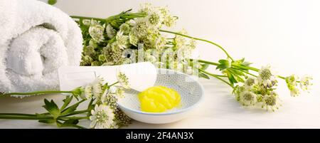 Kosmetische Feuchtigkeitscreme oder Weichmacher und ein Spatel für Pediküre und weiche Fußhaut in einer kleinen Schüssel und einige grüne Blumen auf einem hellen Holzhintergrund, p Stockfoto