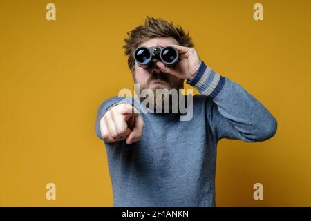 Bizarr zotteliger, bärtiger Mann in einem Strickpullover schaut durch ein Fernglas, er sah etwas und zeigt mit dem Finger. Gelber Hintergrund. Stockfoto