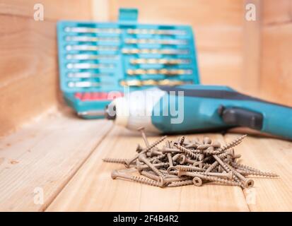 Stapel von Deckschrauben auf Holzbrettern. Viele braune 8x2 Schrauben mit quadratischen Flachköpfen vor dem Elektrobohrer- und Bohrer-Set. Konzept für Out Stockfoto