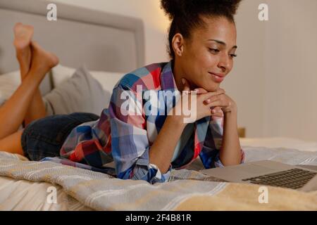 Gemischte Rasse Frau liegt in einem Bett mit einem Laptop Und lächelnd Stockfoto