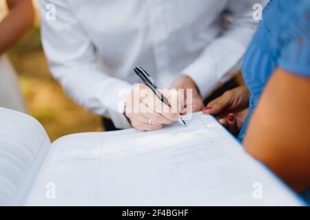 Der Bräutigam unterschreibt während der Hochzeitszeremonie die Hochzeitsurkunde, die Braut steht nahe bei ihm, Nahaufnahme. Stockfoto