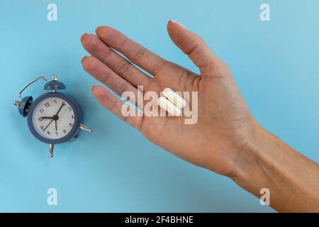 Weiße Kapsel Pille in Frauen Hand auf Wecker Hintergrund. Weibliche Hand mit zwei Kapseln. Zeit bis zur Anwendung der Pille. Stockfoto