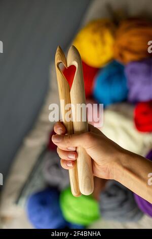 Zwei Holzstricknadeln in einer weiblichen Hand auf dem Hintergrund von mehrfarbigen Merino-Skeins in Form eines Herz Stockfoto