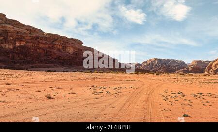 Felsige Massive auf roter Sandwüste, heller wolkiger Himmel im Hintergrund, kleines Fahrzeug und Kamel in der Ferne - typische Landschaft im Wadi Rum, Jordanien Stockfoto