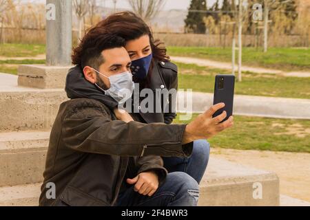 Ein cooles, lächelndes spanisches Paar in Schutzmasken und schwarz Lederjacken, die ein Selfie am Telefon machen Stockfoto