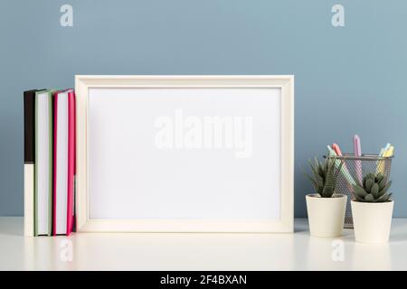 Weißer leerer Holzimitrahmen auf weißem Schreibtisch mit Papierheft, Büchern, Blumen zu Hause Stockfoto