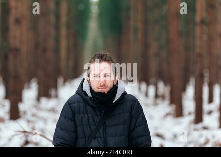 Hübscher junger Mann, der in einem verschneiten Wald steht und gerade aussieht In die Kamera Stockfoto