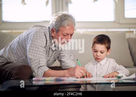 Ein Opa zeigt seinem Enkel, wie man zeichnet, während sie eine gute Zeit in einer entspannten Atmosphäre zu Hause zusammen haben. Familie, Zuhause, Freizeit Stockfoto