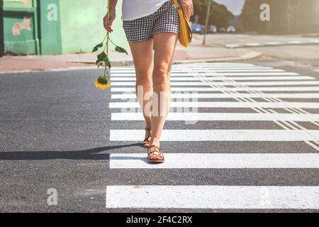 Frau überquert die Straße auf einem Fußgängerüberweg, Sommer in der Stadt. Die Beine der jungen Frau, die Shorts und Sandalen trägt, überqueren die Straße auf Zebra-Markierung. Stockfoto