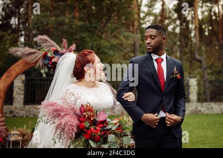Charmanter afroamerikanischer Bräutigam mit wunderschönen weißen Frau stehen in der Nähe dekorierten Bogen, schöne Frau mit schönen Mann auf Hochzeitszeremonie, lächelnd, glücklich Stockfoto