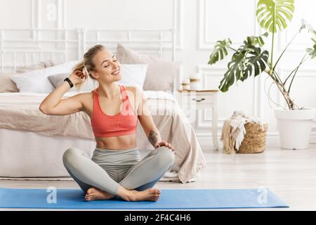 Attraktive blonde Frau trägt Sportkleidung, sitzt auf Gummiteppich, lächelnd, posiert mit geschlossenen Augen. Der junge Yogalehrer ist bereit für die Sitzung Stockfoto