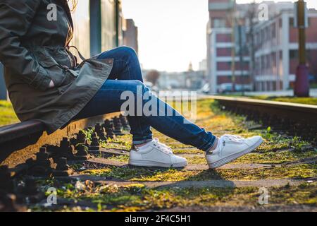 Frau in legerer Kleidung und weißen Sneakers auf Schienen sitzend. Entspannung in der Stadt. Modekonzept Stockfoto