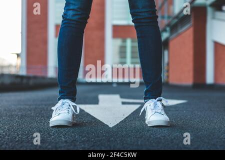 Frau trägt Jeans und weißen Sportschuh stehen auf Asphalt Straße mit Richtungszeichen. Schlanke weibliche Beine mit Sneakers in City Street Stockfoto