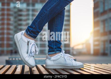 Frau in Jeans und weißen Turnschuhen steht in der Stadt nach Sonnenuntergang. Mode und stilvolles Konzept. Moderne Architektur als Hintergrund Stockfoto
