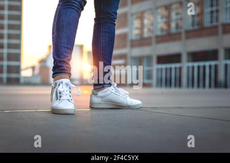 Frau in Jeans und Turnschuhen steht in der Stadt während des Sonnenuntergangs. Selektiver Fokus. Mode und stilvolles Konzept. Weißer Sportschuh im Freien Stockfoto