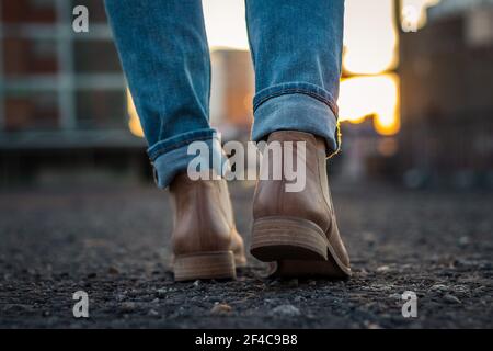 Frau in Jeans und Lederschuhen beim Spaziergang in der Stadt während des Sonnenuntergangs. Nahaufnahme der Schuhe von hinten Stockfoto
