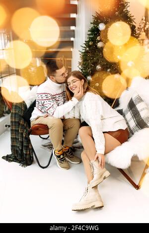 Schönes Paar sitzt in der Nähe von Weihnachtsbaum, lächelnd, charmant Mann sanft küssen liebenswert Frau, glücklich Mann verbringen Ferien mit schönen Frau, gemütlich Stockfoto