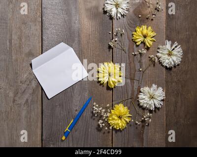 Weiße und gelbe Blüten liegen auf Holzfläche neben Stift und weißem Umschlag. Stockfoto