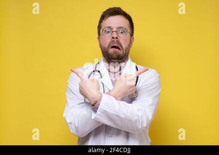 Bizarr, betäubt männlicher Arzt zeigt Zeigefinger in verschiedene Richtungen und sieht mit einem seltsamen Gesichtsausdruck aus. Stockfoto