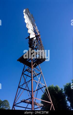 Eine Seitenansicht einer Windmühle, die Wasser aus einer Bohrung zieht, die in vertikalem Format gegen einen blauen Himmel silhouettiert ist. Stockfoto