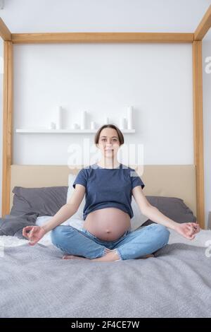 Vorderansicht der jungen erwachsenen schwangeren Frau, die im Bett in Lotusposition sitzt und Meditation praktiziert, Konzept HLS, Yoga Stockfoto