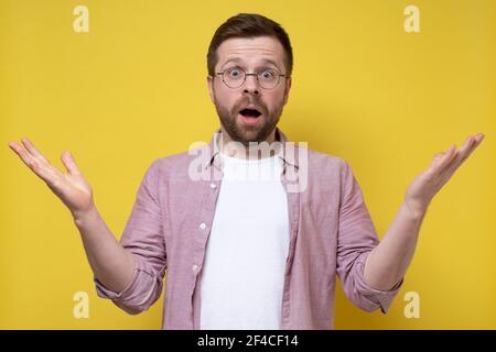 Schockierter Mann in einer Brille und einem Hemd spreizt die Arme an den Seiten und sieht verwirrt aus. Kann nicht sein. Gelber Hintergrund. Stockfoto