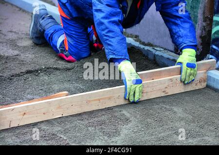 Ein Arbeiter in einem blauen Overalls hockt, um das sandige Fundament mit einer Holzebene zu ebnen, um die Pflasterplatten fortzusetzen, kopieren Raum Bild, selektiver Fokus. Stockfoto