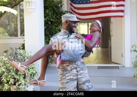 afroamerikanischer Soldat Vater trägt lächelnde Tochter vor Haus Stockfoto