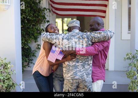 afroamerikanischer Soldat Vater umarmt lächelnde Frau und Vater in Vorderseite des Hauses und amerikanische Flagge Stockfoto