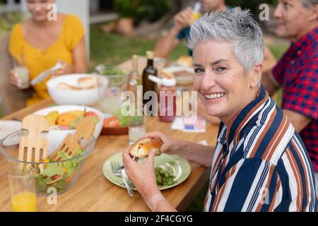 Porträt einer lächelnden kaukasischen älteren Frau, die Hotdog am Tisch hält Mit Familie, die im Garten essen Stockfoto