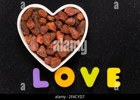 Haufen getrockneter Erdbeeren in einer herzförmigen Schüssel mit Holzblock Buchstaben Schreibweise Liebe. Gesundes Snack-Konzept. Stockfoto