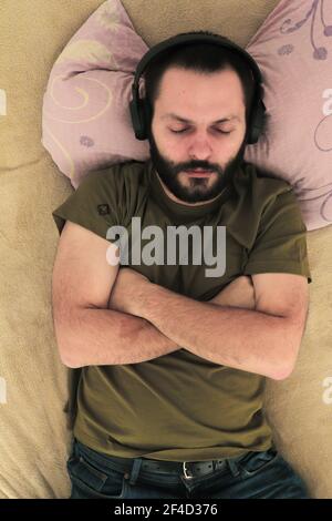 Eine vertikale Aufnahme eines kaukasischen Bartmannes, der lügt Im Bett und Musik über Kopfhörer hören Stockfoto