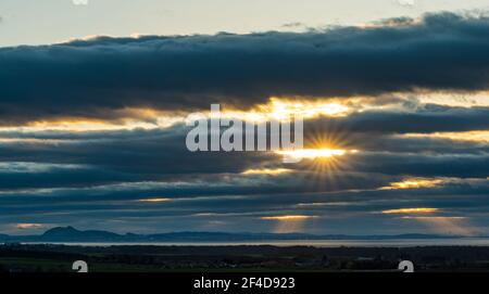 East Lothian, Schottland, Großbritannien, 20th. März 2021. UK Wetter: Sonnenuntergang am Frühlings-Tagundnachtgleiche. Ein wolkenverhallter Himmel über dem markanten Umriss von Edinburgh und Arthur's Sitz ermöglicht einen Ausbruch von Sonnenstrahlen durch, wie die Sonne untergeht Stockfoto