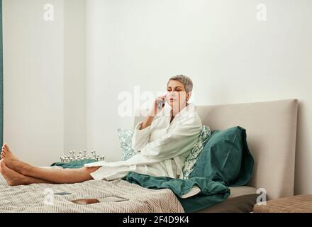 Reife Frau, die am Telefon auf ihrem Bett liegt Stockfoto
