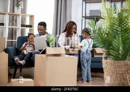 Hübsche afro amerikanische Töchter helfen Eltern zu packen Boxen für Umzug. Junge Familie bereitet sich auf den Umzugstag vor. Besitzer der neuen modernen Wohnung. Stockfoto