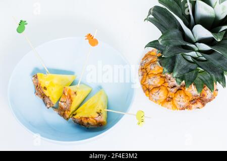 Ananas-Spieße geschält und in Dreiecke mit Essstäbchen verziert geschnitten Im Inneren eine blaue Schale mit weißem Hintergrund und Ananasblättern Stockfoto