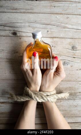 Hände einer jungen Frau mit roten Fingernägeln sind gebunden und liegen auf hellem Holz Hintergrund, hält sie eine kleine Flasche Alkohol, Konzept über Süchtige Stockfoto