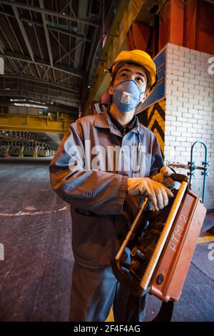 Aluminiumelektrolyseanlage. Der Kranführer für die Schlackenhandhabung mit der Kranfernbedienung. Porträt von jungen asiatischen Arbeiter in gelben Hardhat und blauem Gesicht re Stockfoto