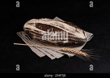 Handgemachtes Sauerteig Roggenbrot auf dunklem Hintergrund. Ganzer Laib Brot auf textiler Serviette und Roggenspikelet Stockfoto