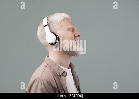 Genießen Sie den Sound. Friedlicher Albino-Mann, der Musik mit geschlossenen Augen im schnurlosen Headset hört und Melodien auf grauem Hintergrund genießt Stockfoto