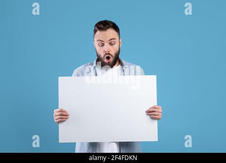 Schockiert bärtigen Mann hält weiße leere Banner auf blauem Studio-Hintergrund, Mockup für Ihre Anzeige oder Design Stockfoto