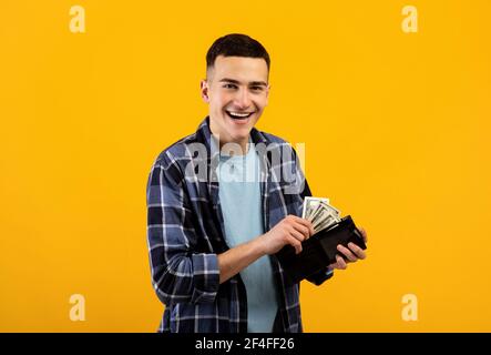 Lächelnder junger Mann, der Geld in seine Geldbörse auf Orange legt studio-Hintergrund Stockfoto