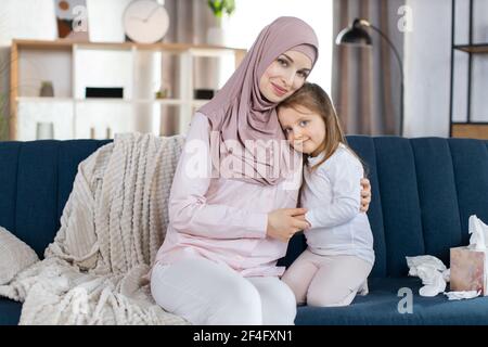 Nahaufnahme Porträt der glücklichen muslimischen Familie, Mutter und kleine Tochter, sitzen zusammen auf blauem Sofa und umarmt, posiert vor der Kamera auf dem Hintergrund der gemütlichen hellen Haus Interieur. Mutterschaft Konzept. Stockfoto