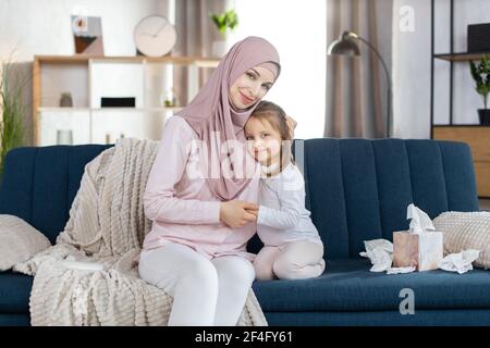 Nahaufnahme Porträt der glücklichen muslimischen Familie, Mutter und kleine Tochter, sitzen zusammen auf blauem Sofa und umarmt, posiert vor der Kamera auf dem Hintergrund der gemütlichen hellen Haus Interieur. Mutterschaft Konzept. Stockfoto