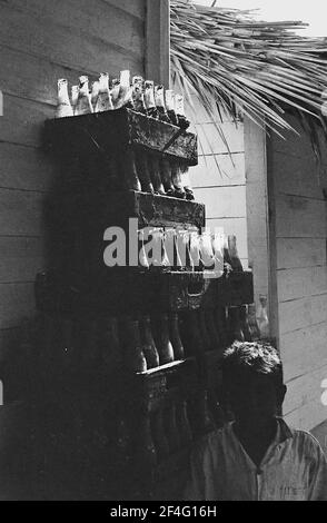 Ein Stapel Coca Cola Flaschen ist unter einem Strohdach sichtbar, mit einer Person in der Ausstellung im Vordergrund in den Sierra Maestra Bergen, Kuba, 1963. Aus der Sammlung Deena Stryker Photographs. () Stockfoto