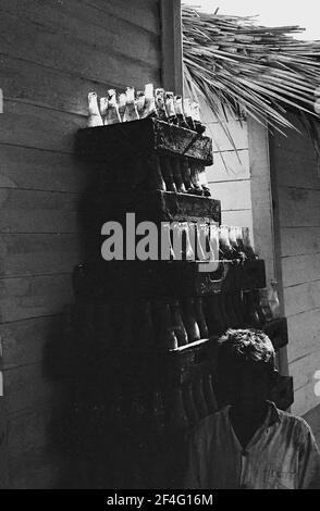 Ein Stapel Coca Cola Flaschen ist unter einem Strohdach sichtbar, mit einer Person in der Ausstellung im Vordergrund in den Sierra Maestra Bergen, Kuba, 1963. Aus der Sammlung Deena Stryker Photographs. () Stockfoto